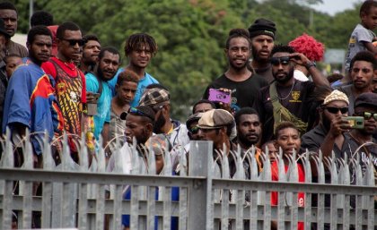 A group of about 16 men from PNG stand watching something over a fence, some with cameras